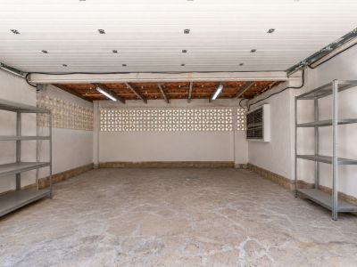 Grand garage avec sol en béton texturé, murs en stuc blanc et blocs de ventilation décoratifs. Avec tuiles en terre cuite apparentes, éclairage fluorescent et étagères métalliques.