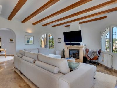 Grand salon avec poutres apparentes en bois, cheminée en pierre et fenêtres cintrées offrant des vues sur le paysage. Canapé modulaire clair et sol en marbre.