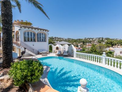 La piscine incurvée aux eaux claires se marie avec la villa méditerranéenne blanche, dotée de fenêtres cintrées et d'une cuisine extérieure avec chaises longues, entourée d'une végétation luxuriante.