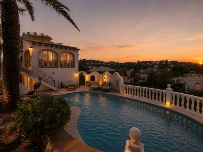 L'extérieur de la villa présente une piscine incurvée et une balustrade éclairée, avec un salon extérieur et des fenêtres cintrées, sous la lueur chaleureuse du coucher de soleil.