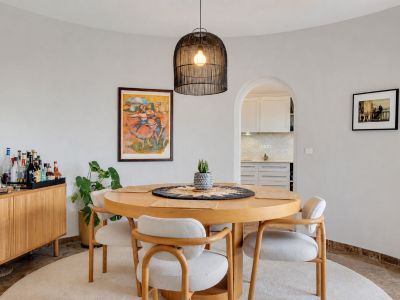 Salle à manger incurvée avec table ronde en bois, quatre chaises rembourrées et une suspension en osier. Une arche mène à la cuisine attenante.