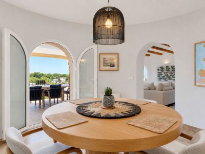 Salle à manger lumineuse avec table ronde en bois et suspension tressée. Des arches mènent à une terrasse extérieure verdoyante et à un salon confortable.