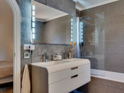Salle de bain contemporaine avec double vasque, meuble incurvé blanc, miroir éclairé par LED et murs en carrelage gris foncé texturé. Une douche intégrée avec paroi en verre est présente.
