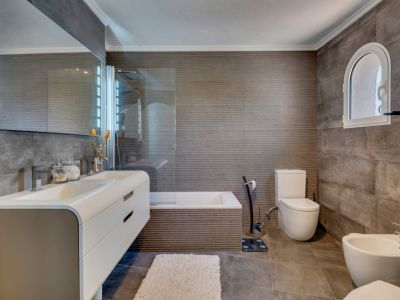 Salle de bain moderne avec carrelage gris texturé, double vasque flottante et miroir éclairé. Une fenêtre cintrée apporte du caractère au-dessus de la zone des toilettes.