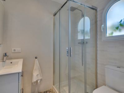 Salle de bain moderne avec une cabine de douche vitrée et pommeau de pluie. Une fenêtre arquée dépolie et un meuble-lavabo blanc complètent cet espace carrelé.