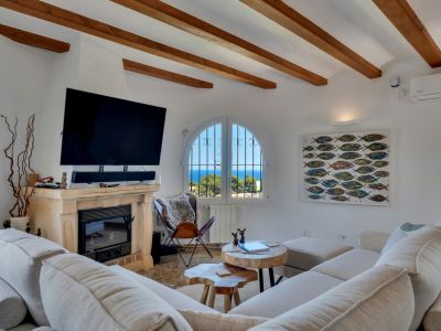 Salon avec poutres en bois apparentes et cheminée en pierre, offrant une vue sur la mer depuis une fenêtre arquée. Canapé modulaire et tables basses en bois naturel.