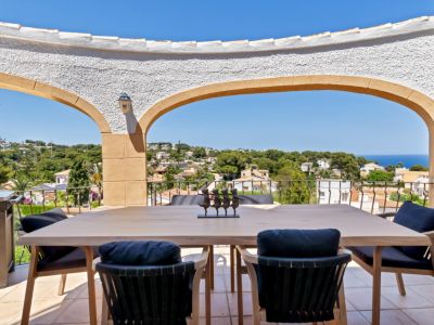 Terrasse couverte avec une table à manger en bois clair et des chaises à coussins sombres, offrant des vues panoramiques sur la mer et la ville à travers des arches méditerranéennes.
