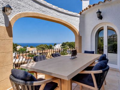 Terrasse repas extérieure avec une table en bois clair et des chaises aux coussins bleus. Une arche encadre des vues panoramiques sur la mer Méditerranée.