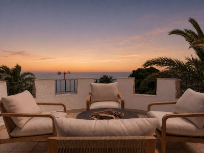 Terrasse sur le toit avec quatre chaises en bois et coussins clairs autour d'un foyer, offrant une vue panoramique sur la mer et des palmiers sous un ciel de coucher de soleil.