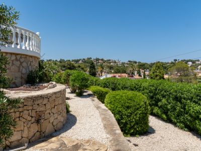 Un mur en pierre sèche incurvé soutient une balustrade blanche, encadrant un chemin de gravier bordé de haies taillées. Des maisons lointaines parsèment la colline verdoyante.