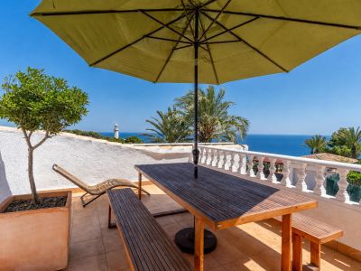 Vaste terrasse avec mobilier de repas en bois sous un parasol, offrant une vue panoramique sur la mer et un phare lointain. Sol carrelé et arbre en pot.