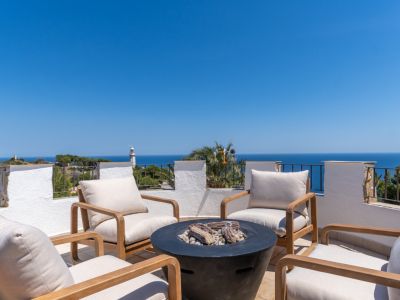 Vaste terrasse sur le toit avec mobilier en bois moderne, foyer central et vues panoramiques sur la mer Méditerranée, incluant un phare lointain.