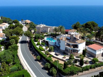 Vue aérienne de la villa méditerranéenne à Jávea, avec piscine surélevée, jardins en terrasses et architecture traditionnelle, face à la vaste mer bleue et un phare lointain.