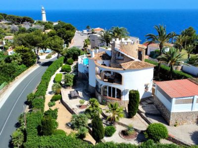 Vue aérienne d'une villa méditerranéenne avec murs blancs, tuiles et piscine. Des jardins luxuriants entourent la propriété, avec la mer et un phare en arrière-plan.