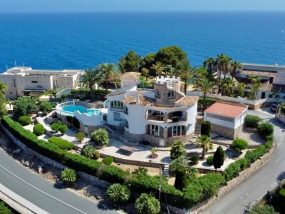 Vue aérienne d'une villa méditerranéenne avec tour ronde distinctive, jardins en terrasses et piscine, le tout face à la vaste mer bleue.