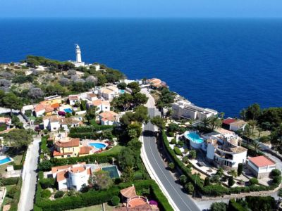Vue aérienne panoramique d'une urbanisation côtière avec villas, piscines privées et jardins luxuriants, bordant la mer Méditerranée d'un bleu profond et un phare lointain.