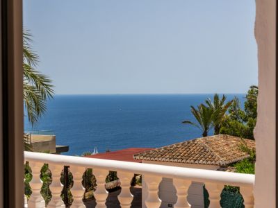 Vue depuis un balcon avec balustrade blanche, toit en terre cuite et palmiers luxuriants, surplombant la vaste mer Méditerranée avec un voilier au loin.