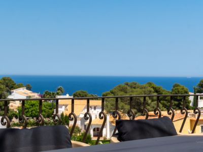 Vue depuis une terrasse avec balustrade en fer forgé, surplombant des toits en terre cuite, des pins et la mer Méditerranée.