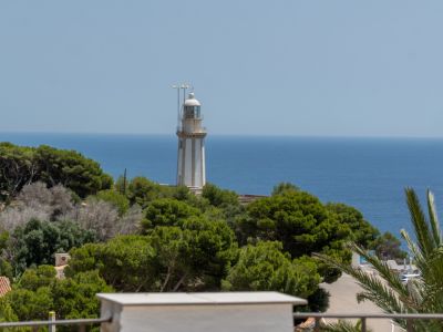 Vue du phare blanc rayé de gris, dressé sur une colline verdoyante, avec la vaste mer Méditerranée bleue s'étendant à l'horizon sous un ciel clair.