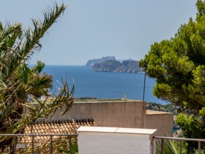 Vue panoramique sur la mer avec une côte rocheuse lointaine, encadrée par des palmiers et des pins. Des tuiles de terre cuite traditionnelles sont visibles en contrebas.