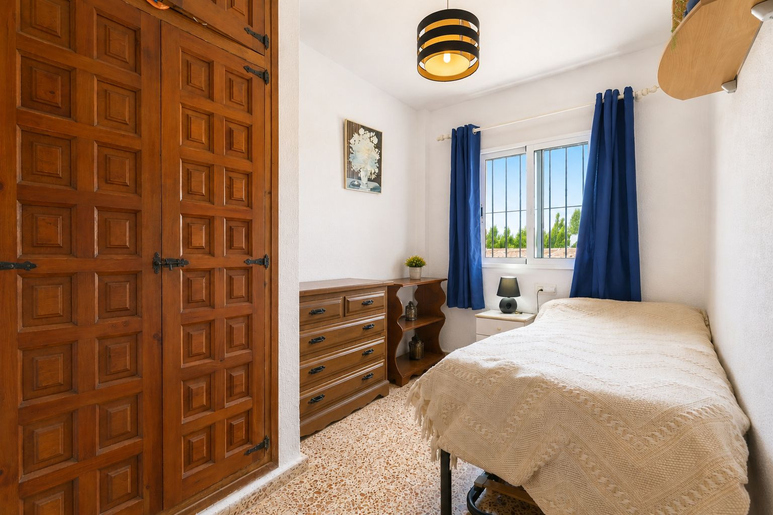Chambre confortable avec armoire encastrée en bois à panneaux décoratifs et ferrures noires. Fenêtre avec rideaux bleus et vue sur la verdure, apportant lumière naturelle.