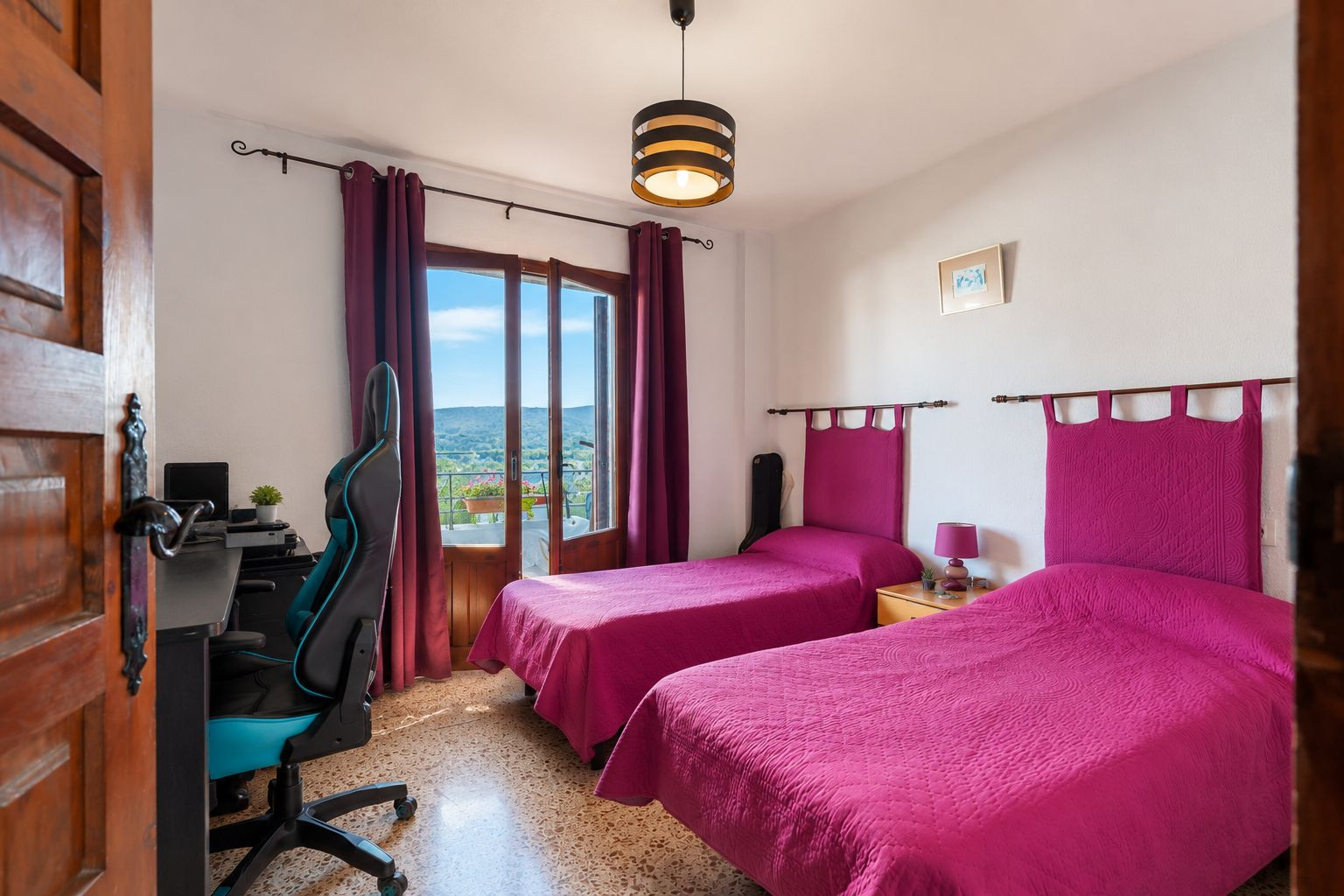 Chambre lumineuse avec deux lits simples, têtes de lit fuchsia et accès direct à un balcon avec vue sur la montagne. Comprend un coin bureau et un sol en terrazzo.