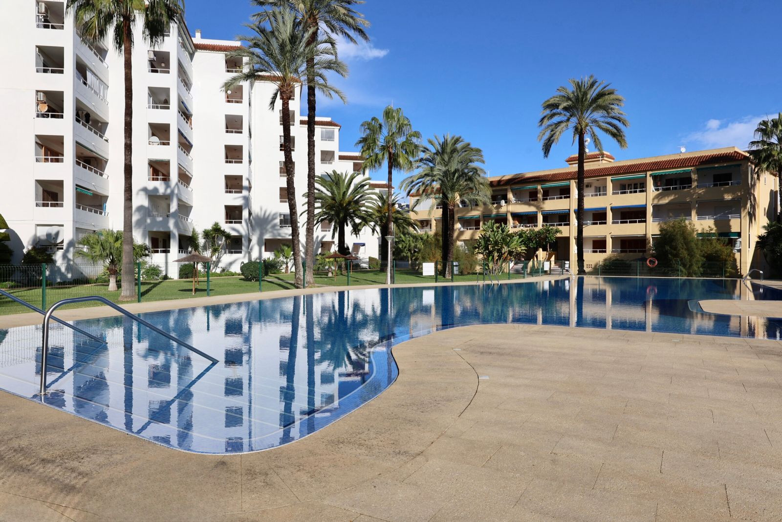 La grande piscine commune, avec ses carreaux bleus et sa plage carrelée claire, est dotée de rampes en acier. Palmiers et bâtiments résidentiels se reflètent dans son eau sous un ciel bleu.