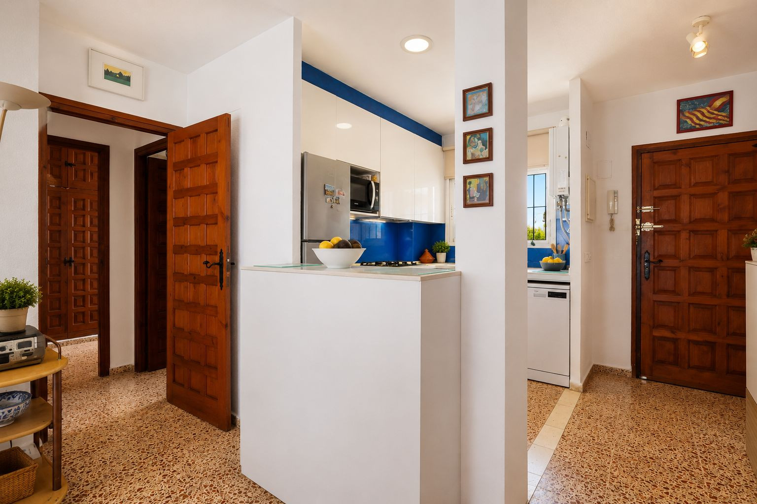 Le hall d'entrée présente un carrelage à motifs distinctifs et des portes en bois classiques. Une cuisine moderne avec crédence bleue et meubles blancs est visible, séparée par un comptoir fonctionnel.