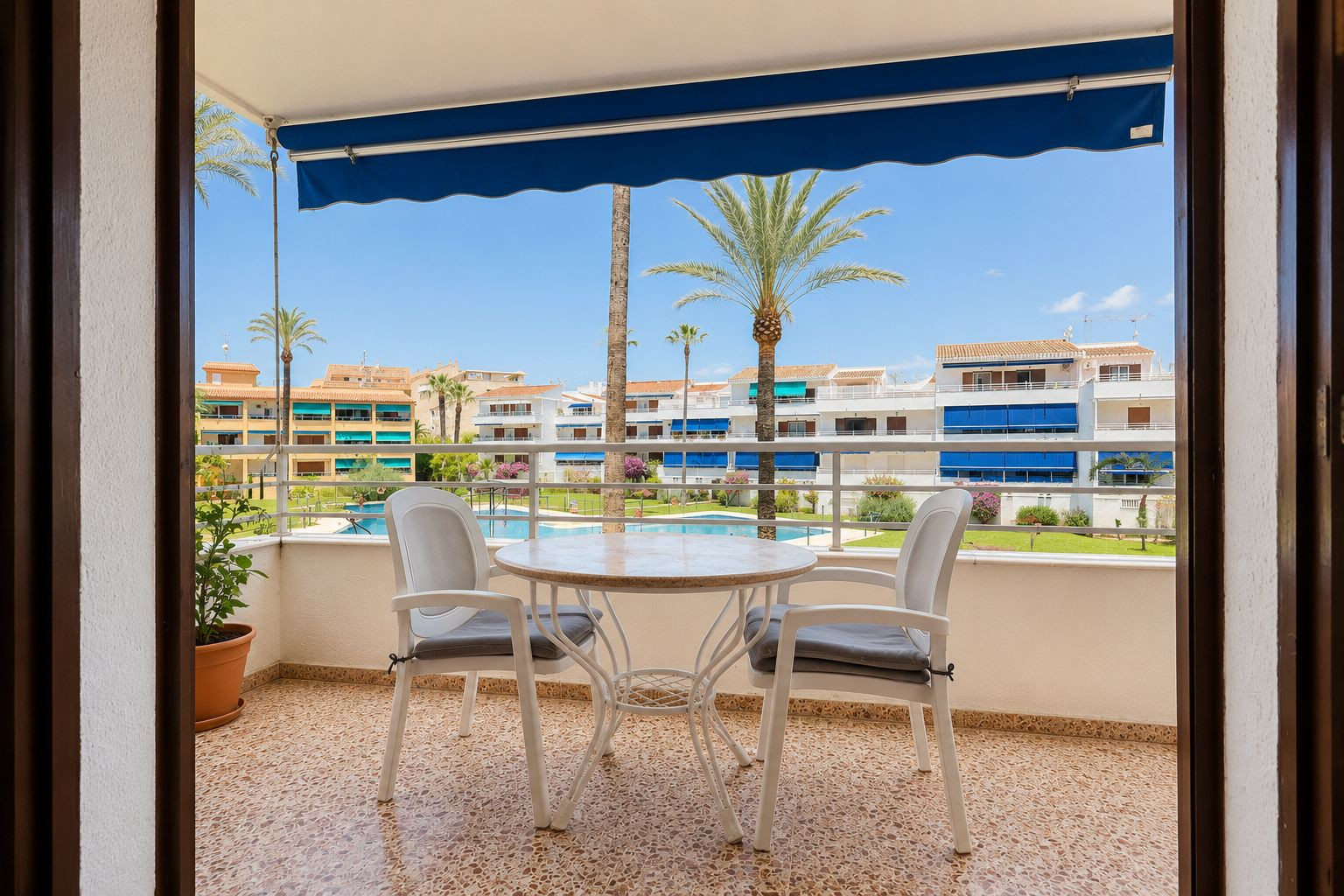 Terrasse carrelée avec une table ronde et deux chaises, protégée par un store bleu. Elle offre une vue sur la piscine commune, les jardins et les palmiers.