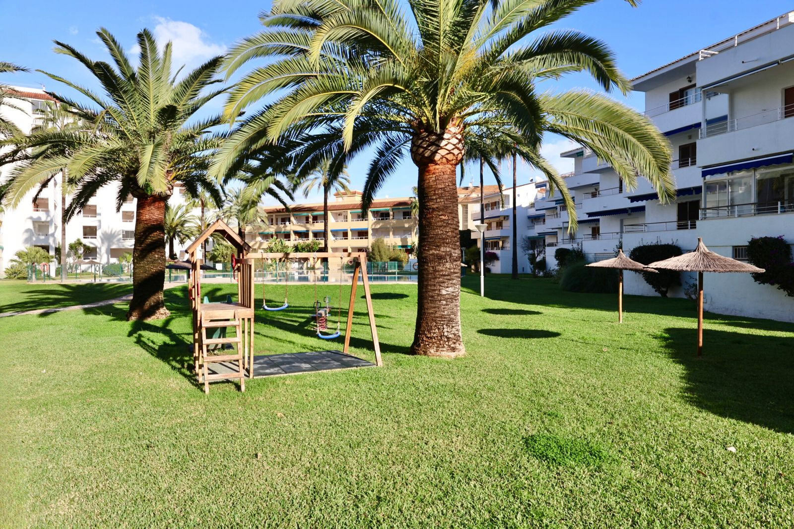 Vaste jardin commun avec pelouse verdoyante, aire de jeux pour enfants en bois, palmiers matures et deux parasols en paille, devant des résidences.