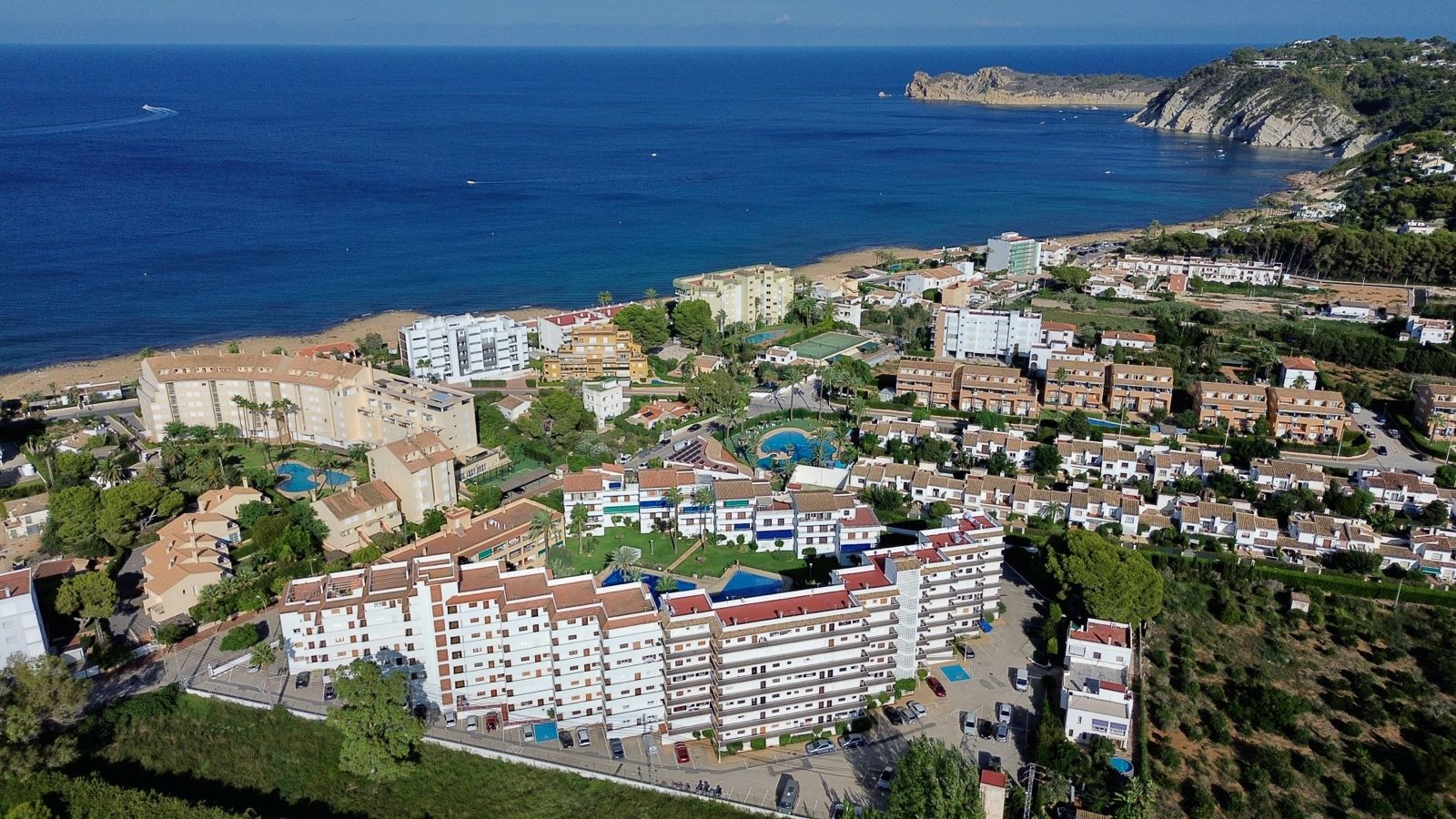 Vue aérienne d'une urbanisation côtière avec plusieurs bâtiments résidentiels, piscines communes et espaces verts, bordant une plage de sable et la mer Méditerranée, avec un cap rocheux au loin.