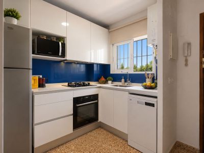 Cuisine moderne et fonctionnelle avec des armoires blanches brillantes, un plan de travail clair et une crédence bleue. Elle est équipée d'appareils intégrés et d'une fenêtre avec vue sur la verdure.