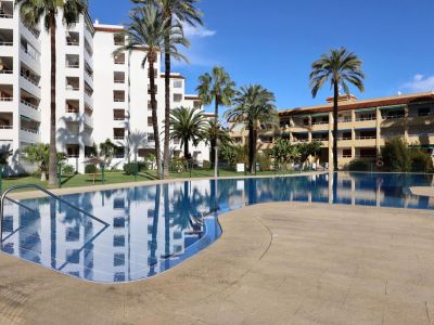 La grande piscine commune, avec ses carreaux bleus et sa plage carrelée claire, est dotée de rampes en acier. Palmiers et bâtiments résidentiels se reflètent dans son eau sous un ciel bleu.