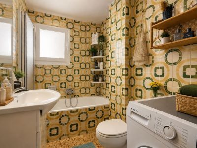 La salle de bain complète arbore un carrelage distinctif à motifs géométriques jaunes et verts, un lavabo avec meuble, une baignoire avec pare-douche et une machine à laver intégrée, éclairée par une fenêtre dépolie.