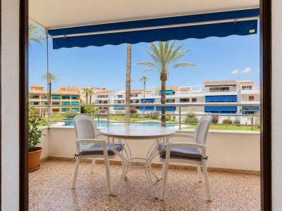 Terrasse carrelée avec une table ronde et deux chaises, protégée par un store bleu. Elle offre une vue sur la piscine commune, les jardins et les palmiers.