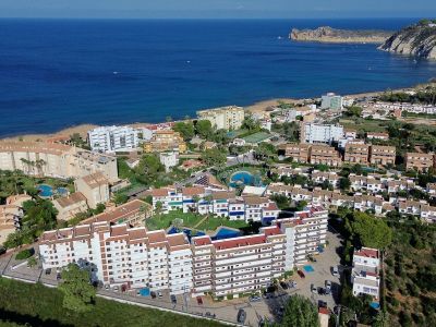 Vue aérienne d'une urbanisation côtière avec plusieurs bâtiments résidentiels, piscines communes et espaces verts, bordant une plage de sable et la mer Méditerranée, avec un cap rocheux au loin.