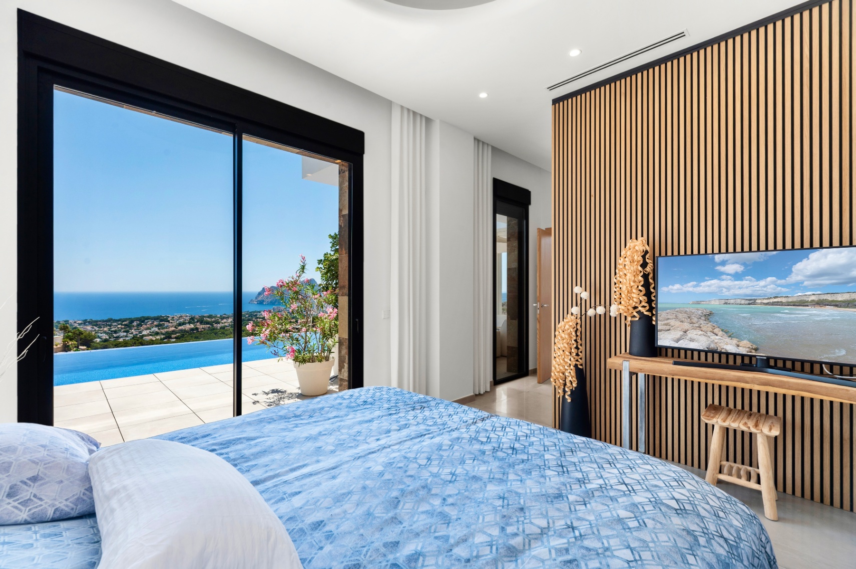 Chambre avec de grandes portes coulissantes en verre s'ouvrant sur une piscine à débordement et des vues panoramiques sur la mer. Un mur moderne en lattes de bois encadre la télévision, complétant l'intérieur clair. Chambre avec de grandes portes coulissantes en verre s'ouvrant sur une piscine à débordement et des vues panoramiques sur la mer. Un mur moderne en lattes de bois encadre la télévision, complétant l'intérieur clair.