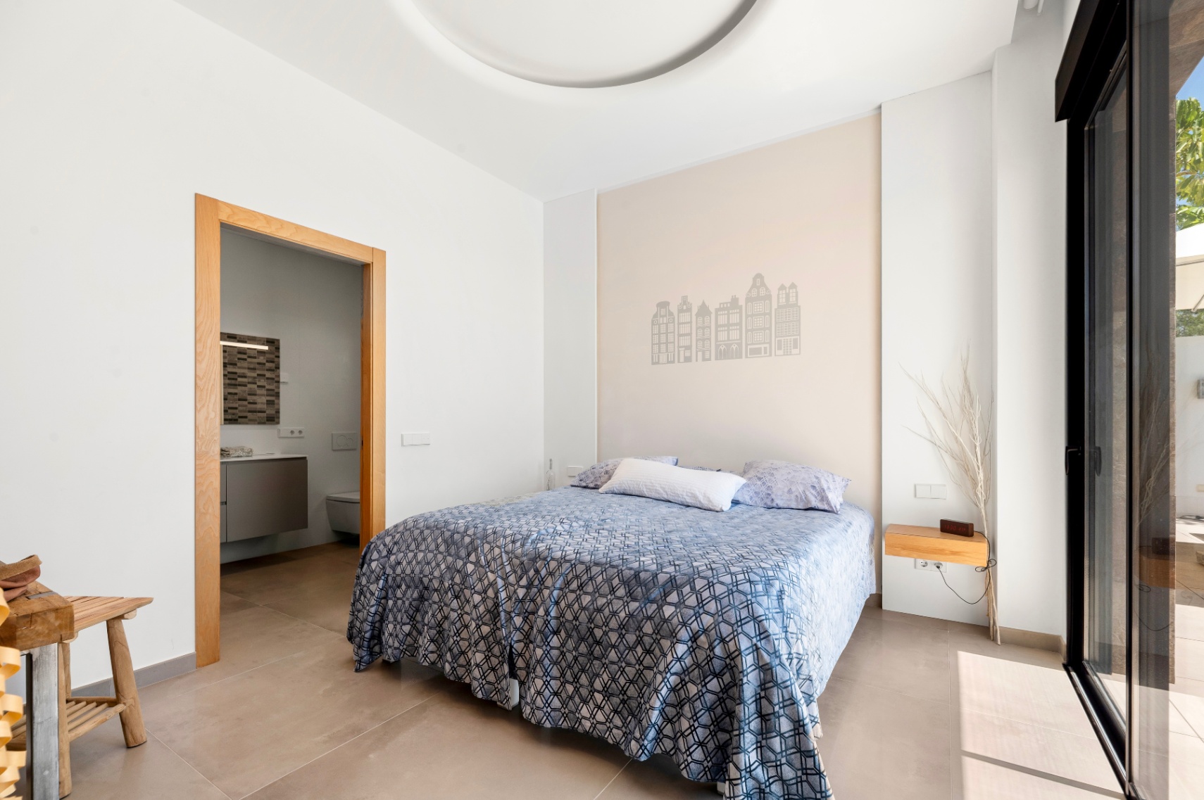 Chambre lumineuse avec lit double, un mur d'accent décoré de silhouettes de bâtiments et accès à une salle de bain attenante. Porte coulissante vitrée menant directement à une terrasse extérieure. Chambre lumineuse avec lit double, un mur d'accent décoré de silhouettes de bâtiments et accès à une salle de bain attenante. Porte coulissante vitrée menant directement à une terrasse extérieure.