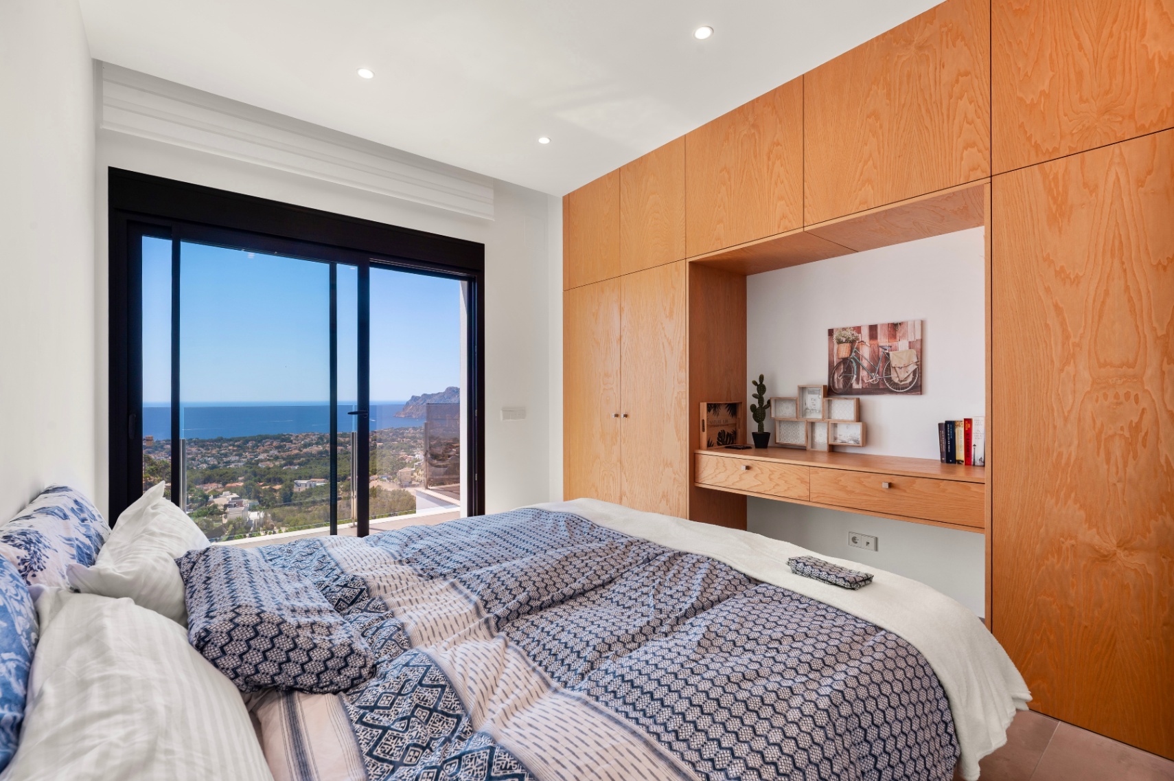 Chambre principale avec de grandes baies vitrées coulissantes offrant une vue panoramique sur la mer et la côte. Armoires intégrées en bois clair avec bureau. Chambre principale avec de grandes baies vitrées coulissantes offrant une vue panoramique sur la mer et la côte. Armoires intégrées en bois clair avec bureau.