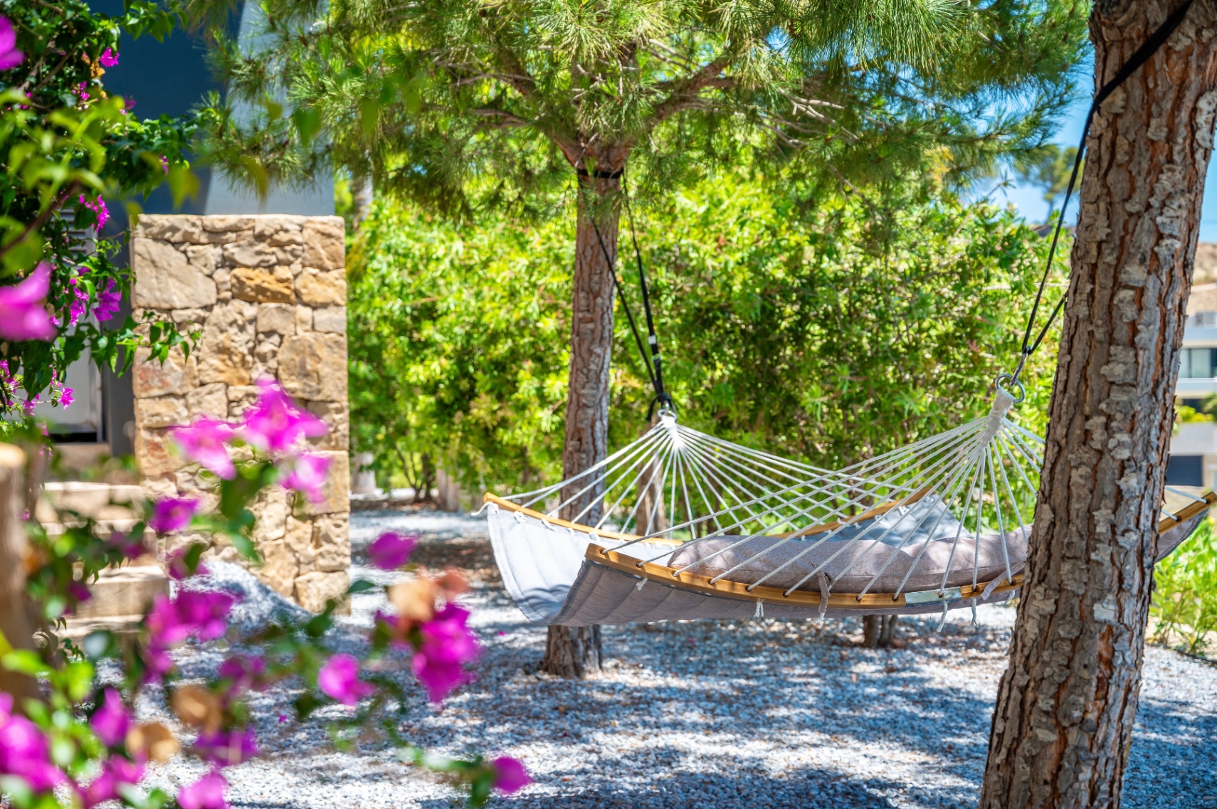 Coin de jardin paisible avec un hamac gris confortable à barres en bois, suspendu entre des arbres sur un lit de gravier, encadré par des bougainvilliers éclatants. Coin de jardin paisible avec un hamac gris confortable à barres en bois, suspendu entre des arbres sur un lit de gravier, encadré par des bougainvilliers éclatants.