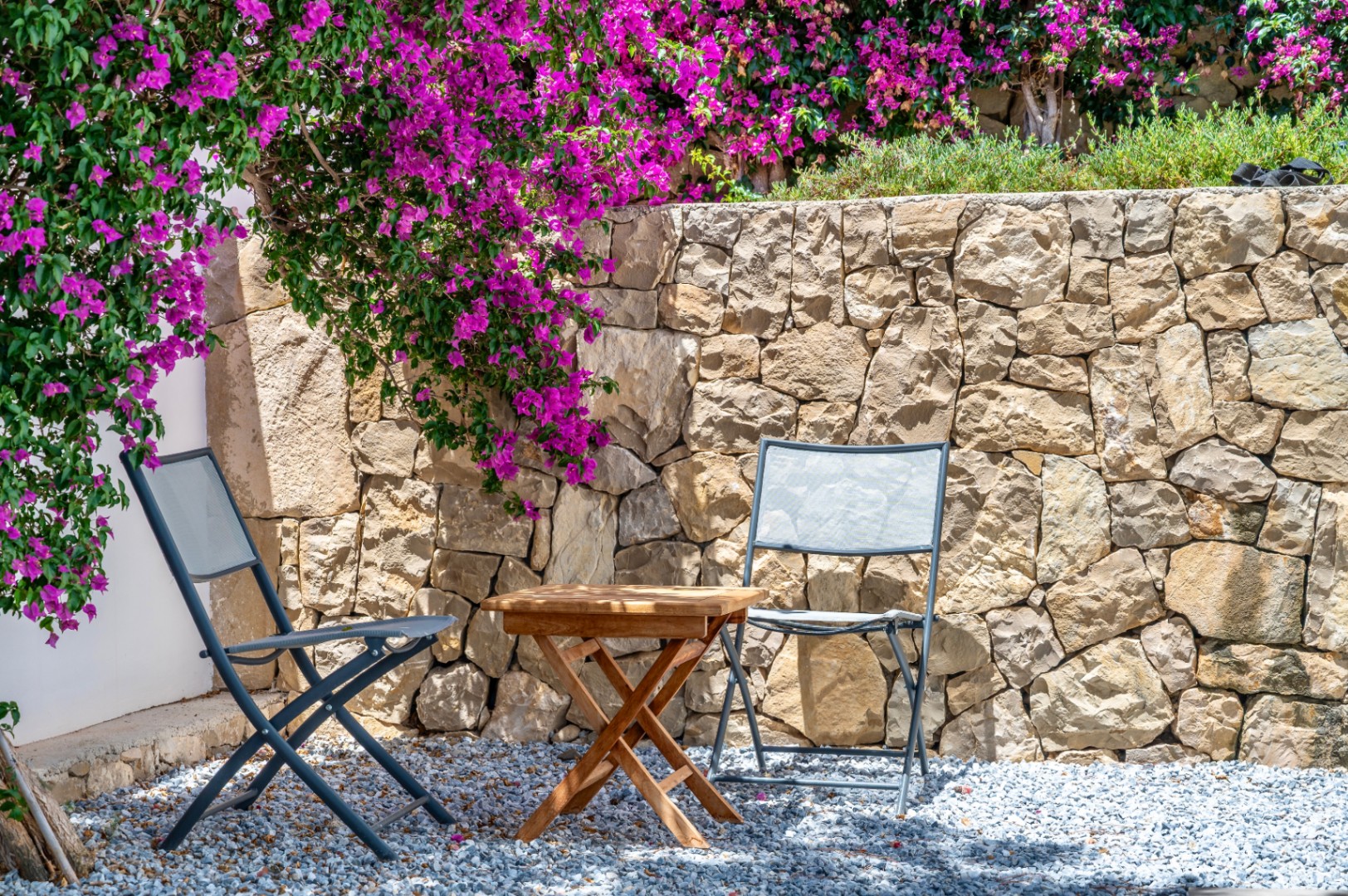 Coin salon extérieur avec deux chaises pliantes grises et une table en bois sur gravier. Un mur en pierre naturelle est orné de bougainvilliers magenta éclatants. Coin salon extérieur avec deux chaises pliantes grises et une table en bois sur gravier. Un mur en pierre naturelle est orné de bougainvilliers magenta éclatants.