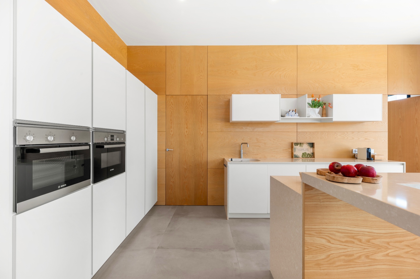 Cuisine moderne avec armoires blanches intégrées et fours Bosch. L'îlot central en pierre claire et les panneaux muraux et plafond en bois clair caractérisent l'espace. Cuisine moderne avec armoires blanches intégrées et fours Bosch. L'îlot central en pierre claire et les panneaux muraux et plafond en bois clair caractérisent l'espace.