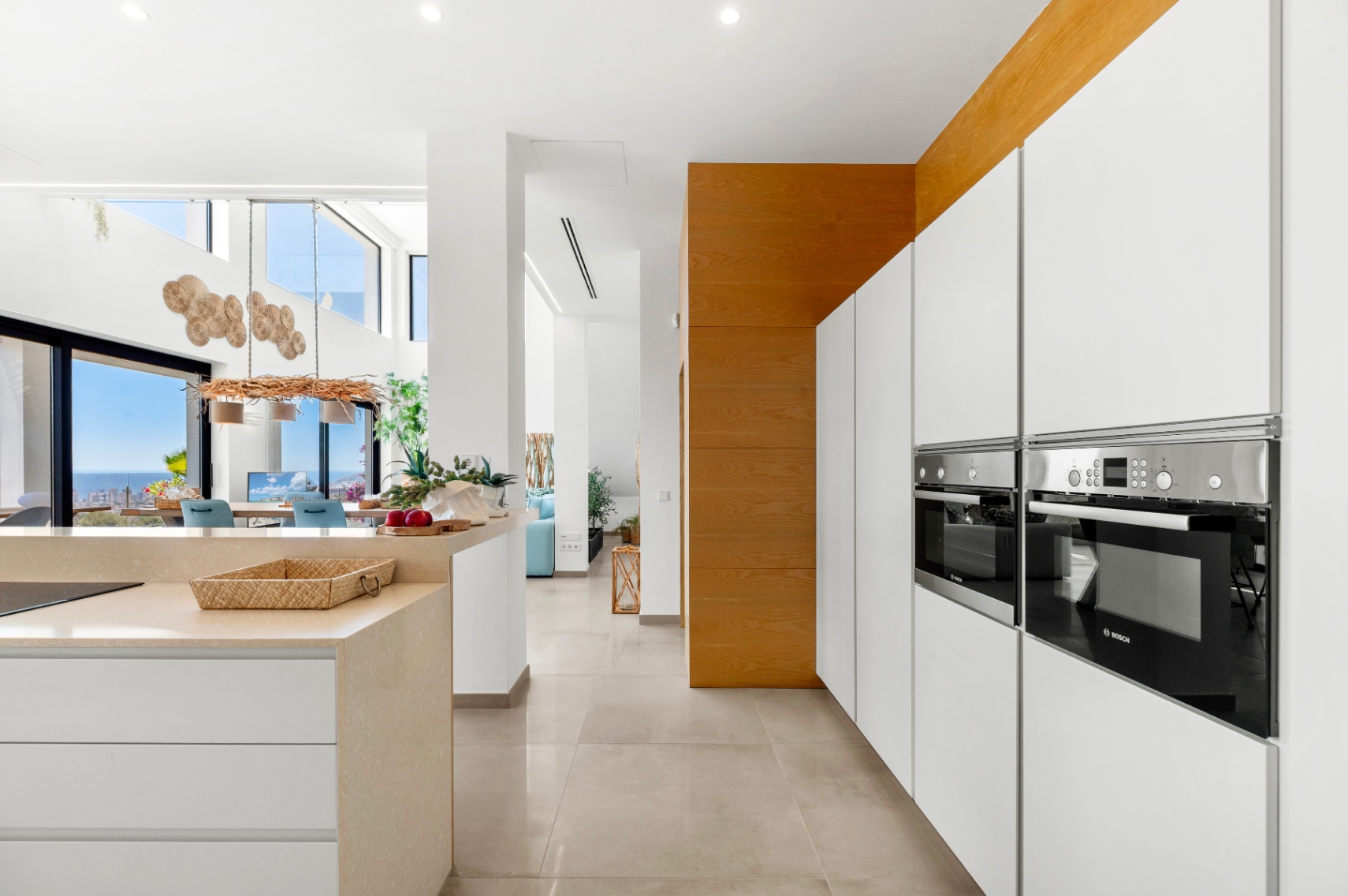 Cuisine moderne épurée avec fours Bosch intégrés, armoires blanches sans poignées et îlot en pierre claire. L'agencement ouvert s'étend vers la salle à manger avec vue panoramique sur la mer. Cuisine moderne épurée avec fours Bosch intégrés, armoires blanches sans poignées et îlot en pierre claire. L'agencement ouvert s'étend vers la salle à manger avec vue panoramique sur la mer.