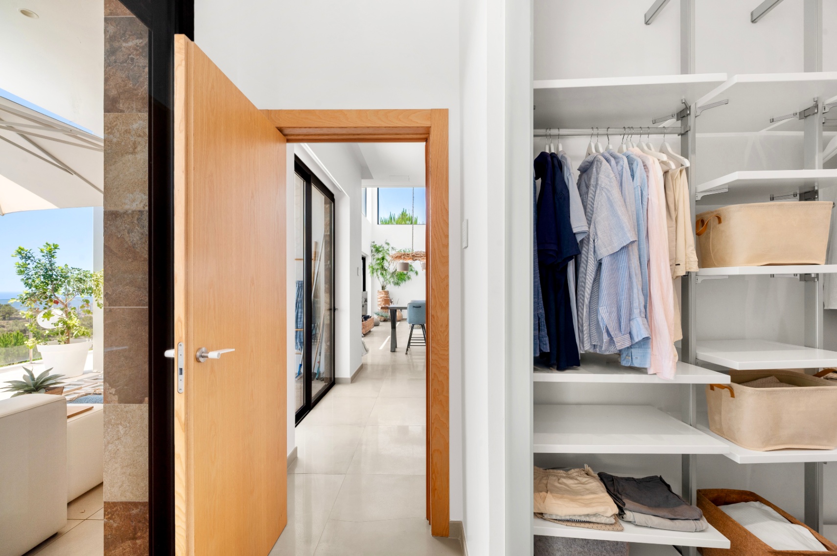 Entrée de couloir moderne avec porte en bois clair ouverte, révélant un placard blanc intégré avec étagères modulables et un aperçu de terrasse ensoleillée avec vue sur mer. Entrée de couloir moderne avec porte en bois clair ouverte, révélant un placard blanc intégré avec étagères modulables et un aperçu de terrasse ensoleillée avec vue sur mer.