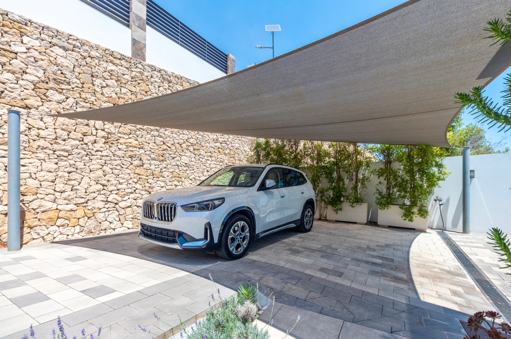 Espace de stationnement extérieur abrité par une voile d'ombrage, avec une allée pavée bicolore. Un mur en pierres sèches et des jardinières végétalisées encadrent la zone. Espace de stationnement extérieur abrité par une voile d'ombrage, avec une allée pavée bicolore. Un mur en pierres sèches et des jardinières végétalisées encadrent la zone.