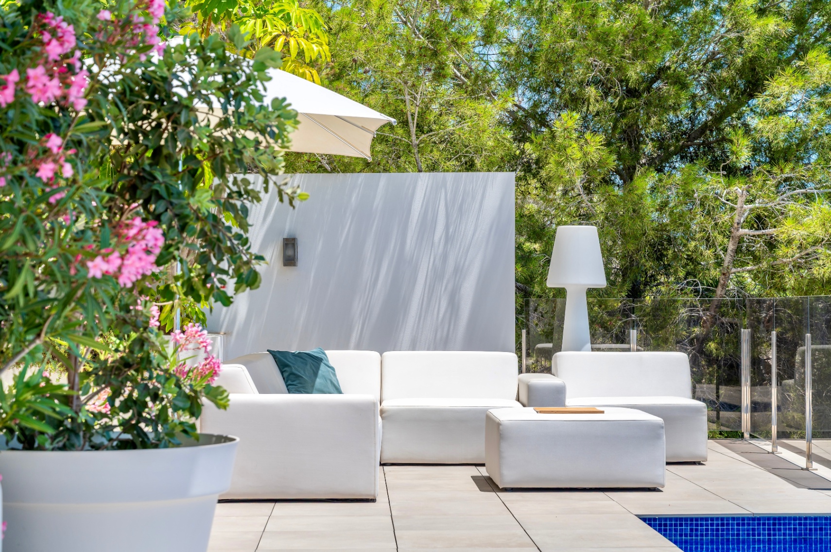 Espace lounge extérieur contemporain avec canapés modulaires blancs et pouf assorti sur une terrasse carrelée. Bordé par une rambarde en verre, une végétation luxuriante et une plante à fleurs roses. Espace lounge extérieur contemporain avec canapés modulaires blancs et pouf assorti sur une terrasse carrelée. Bordé par une rambarde en verre, une végétation luxuriante et une plante à fleurs roses.