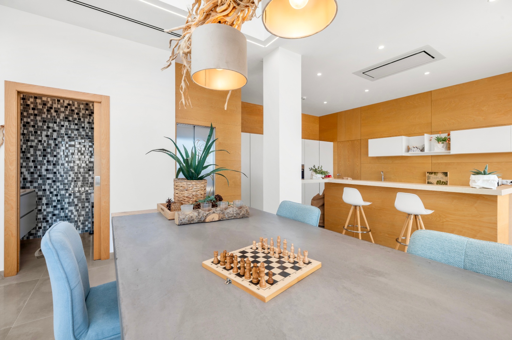 Espace salle à manger contemporain avec table grise et chaises bleues. Un lustre en bois naturel et une cuisine ouverte avec boiseries claires et comptoir sont visibles. Accès à une salle de bain carrelée. Espace salle à manger contemporain avec table grise et chaises bleues. Un lustre en bois naturel et une cuisine ouverte avec boiseries claires et comptoir sont visibles. Accès à une salle de bain carrelée.
