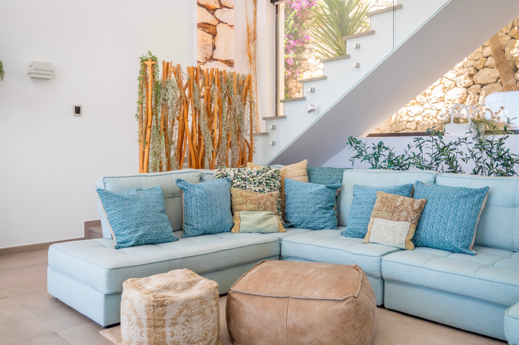 Espace salon chaleureux avec canapé sectionnel bleu clair, coussins variés et deux poufs en cuir. Escalier moderne en verre près d'un écran en bois naturel et plantes, mur de pierre visible. Espace salon chaleureux avec canapé sectionnel bleu clair, coussins variés et deux poufs en cuir. Escalier moderne en verre près d'un écran en bois naturel et plantes, mur de pierre visible.