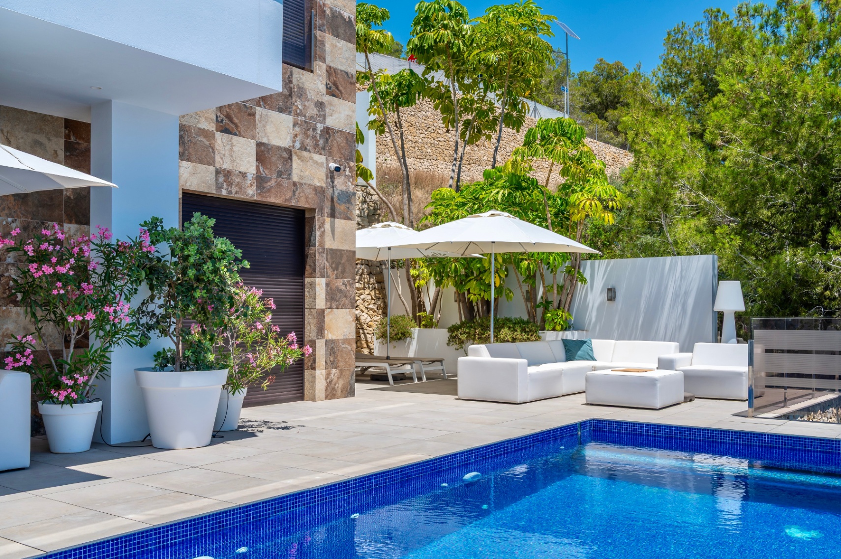 Grande terrasse avec piscine en mosaïque bleue et mobilier extérieur blanc sous parasols, agrémentée d'une façade en pierre et d'une végétation méditerranéenne luxuriante. Grande terrasse avec piscine en mosaïque bleue et mobilier extérieur blanc sous parasols, agrémentée d'une façade en pierre et d'une végétation méditerranéenne luxuriante.