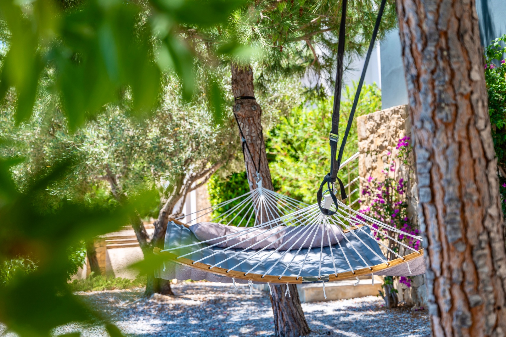 Hamac gris rembourré avec coussin, suspendu entre des arbres feuillus sur un sol de gravier. Un mur en pierre avec des fleurs violettes orne l'arrière-plan du jardin. Hamac gris rembourré avec coussin, suspendu entre des arbres feuillus sur un sol de gravier. Un mur en pierre avec des fleurs violettes orne l'arrière-plan du jardin.